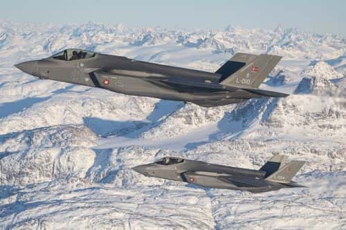 To danske F-35-kampfly flyver i formation over et snedækket bjerglandskab langs Grønlands østkyst. Billedet illustrerer Danmarks styrkede overvågning og militære tilstedeværelse i Arktis og Nordatlanten.