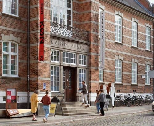 Indgang til museet KØN (tidl. Kvindemuseet) i Aarhus i en rød murstensbygning. Skiltet “KVINDEMUSEET” står over døren, og bannere med “KØN” hænger på facaden. Flere personer går op ad trappen, og cykler står langs bygningen.