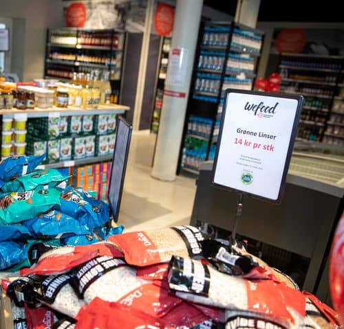 Hylder og varer i en Wefood-butik. I forgrunden ligger poser med tørrede bælgfrugter, blandt andet grønne linser. Et skilt viser teksten "Grønne Linser – 14 kr pr stk". I baggrunden ses kølediske og butikkens vareudvalg.