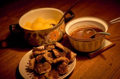 Tallerken med stegte frikadeller på et spisebord ved siden af en gryde med kartofler og en kasserolle med brun sovs. Retten er et eksempel på dansk madkultur og hverdagslige traditioner, der kan gå i arv mellem generationer.