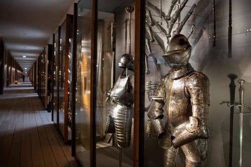 Interiør fra Krigsmuseet med en lang gang, hvor rustninger og våben er udstillet i glasmontrer langs væggen. En blank metalrustning står fremtrædende i forgrunden, mens flere andre rustninger og sværd ses i montrerne bagved.