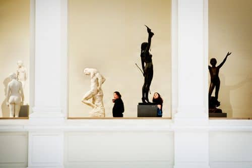 Interiør fra SMK med flere klassiske skulpturer placeret i nicher mellem hvide søjler; både lyse marmorskulpturer og en mørk bronzestatue med armen løftet. To besøgende står i forgrunden og kigger på værkerne.