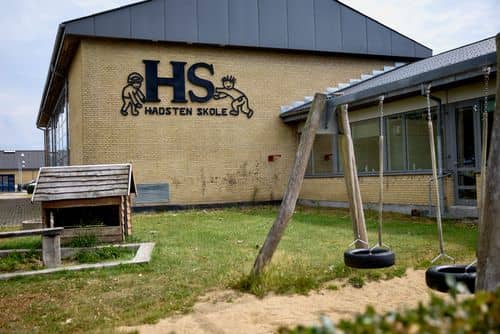 Bygning med skiltet "HS Hadsten Skole" samt skolegård med gynger og legeområde foran skolens facade.