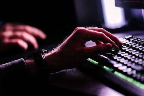 xxNærbillede af hænder ved et computerkeyboard i et mørkt rum med lys fra en skærm. Billedet illustrerer digital aktivitet og kommunikation online.