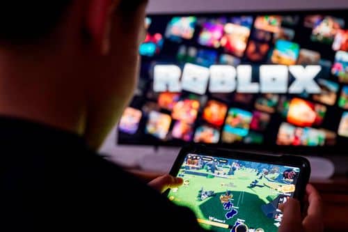 Person holder en mobiltelefon med Roblox åbent, mens Roblox-logoet og forskellige spiluniverser vises på en skærm i baggrunden.