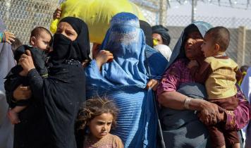 Tildækkede afghanske kvinder med børn på armen står foran et højt pigtrådshegn.