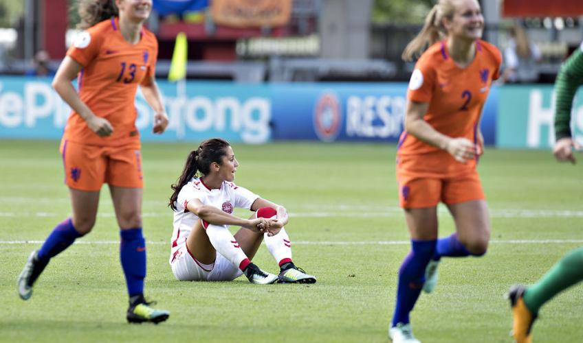 Den danske landsholdsspiller Nadia Nadim sidder på græsset, mens hollandske spillere løber glade rundt.