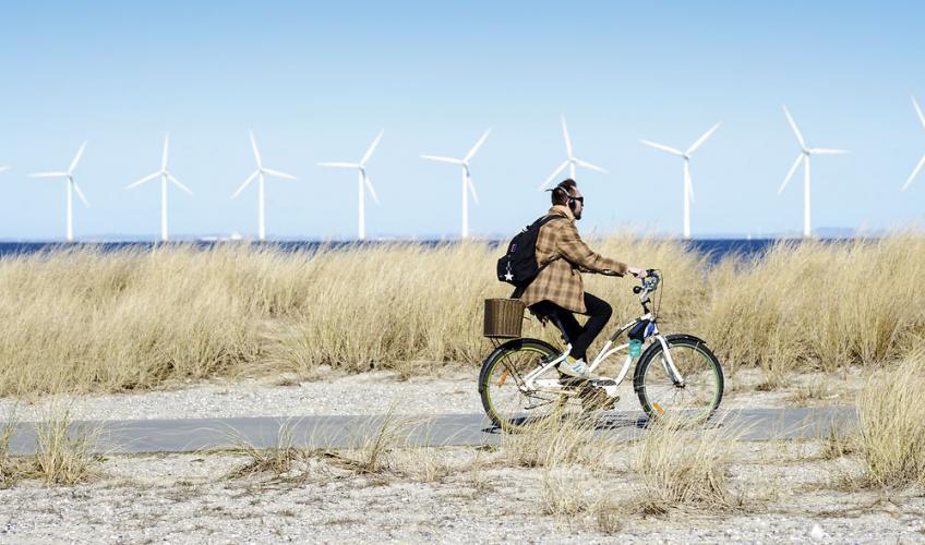 En mand cykler  med vindmøller ude i havet bag ham.